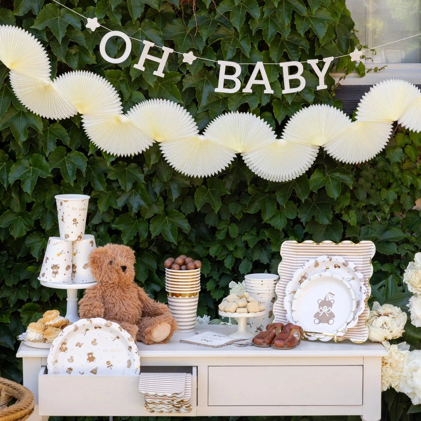 Baby shower setup with 'OH BABY' banner, teddy bear plates, and decorations against a green leafy background.