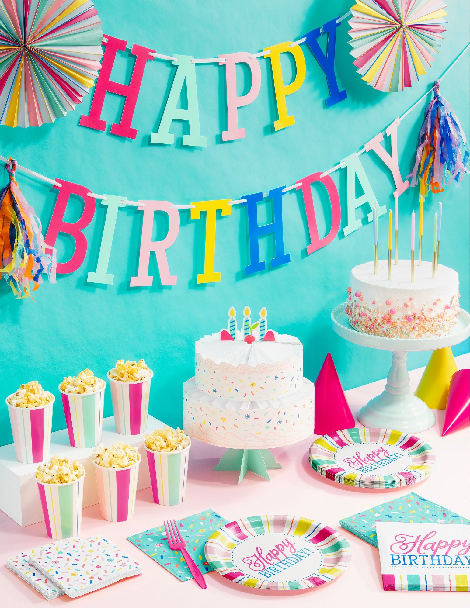 Birthday party setup with cake, decorations, and tableware against a turquoise background