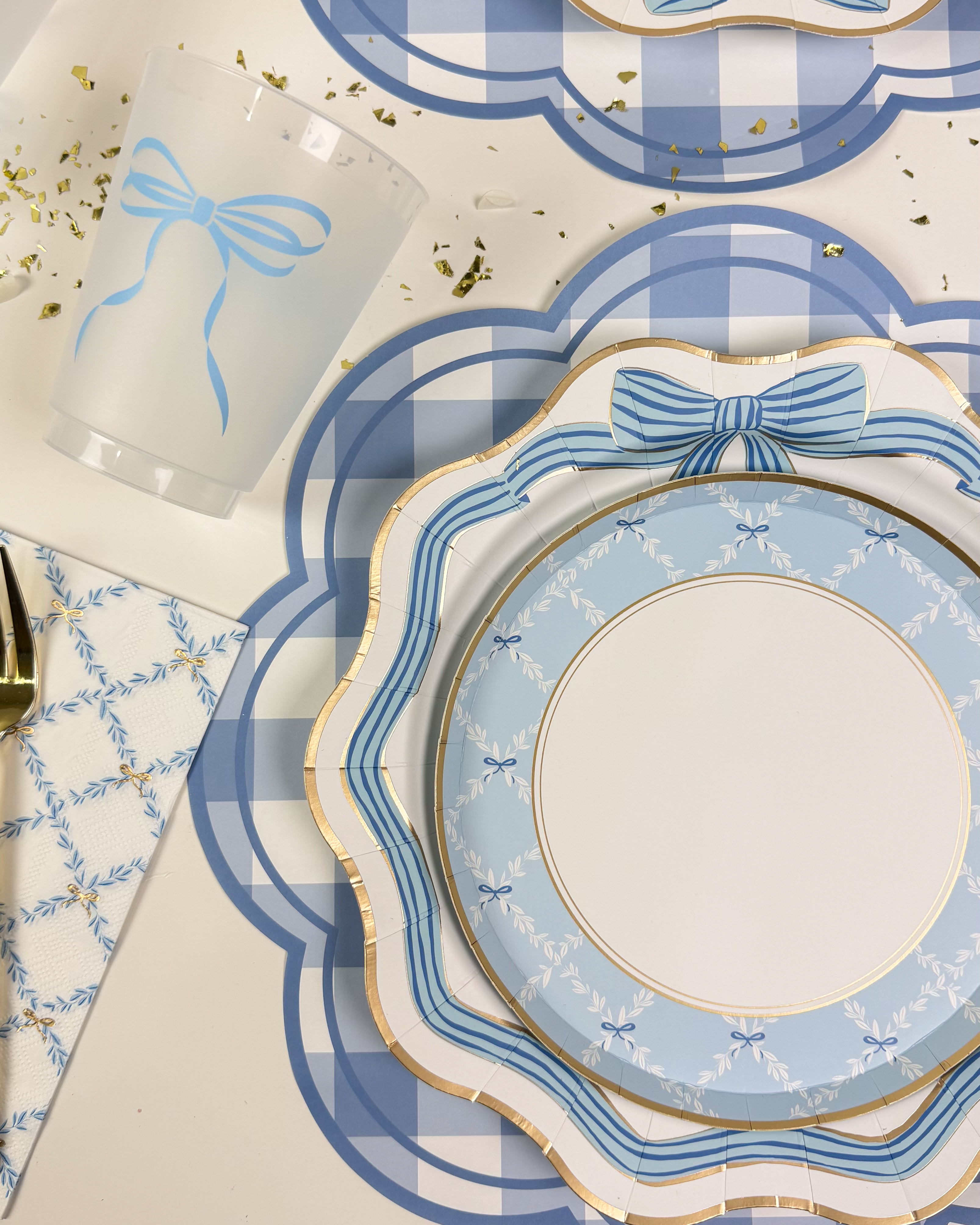Overhead view of a blue and white bow-themed table setting with gold-trimmed plates, frosted bow cups, napkins, and gold confetti.