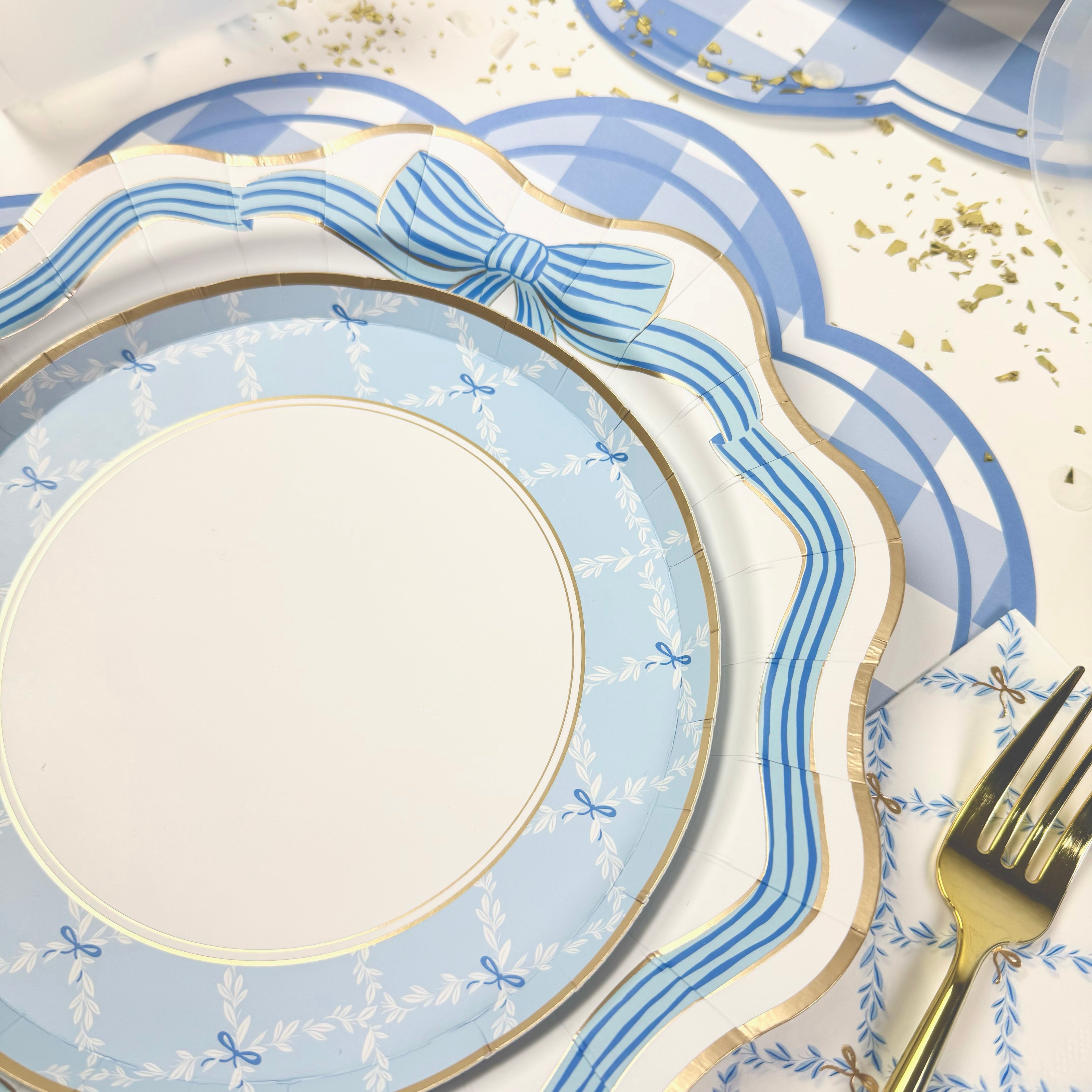 Blue scalloped paper plates with gold trim and bow detail, styled with a gold fork, patterned napkin, and gingham placemats.