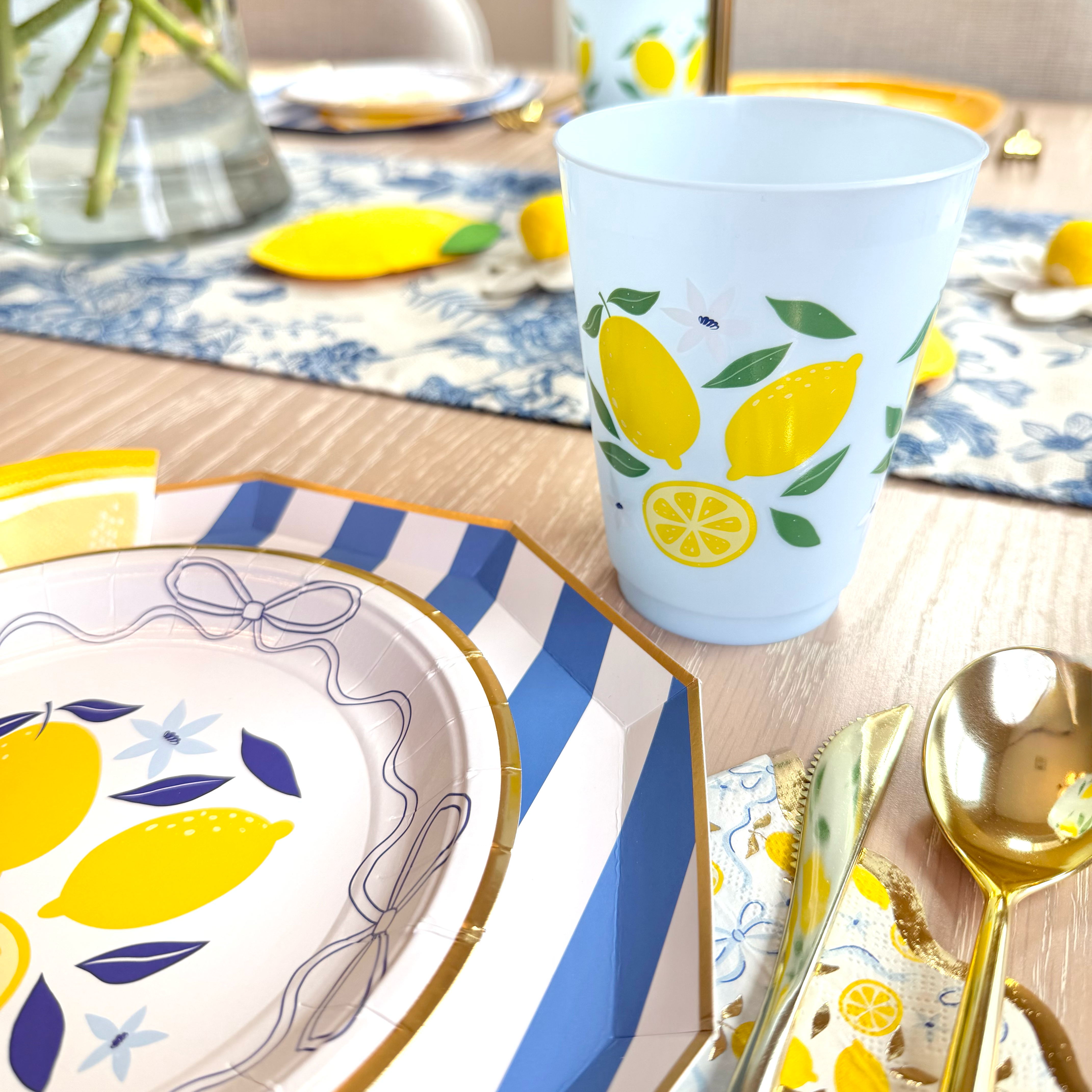 Close-up of lemon party place setting with blue striped charger, lemon plate, lemon-print cup, and gold utensils.