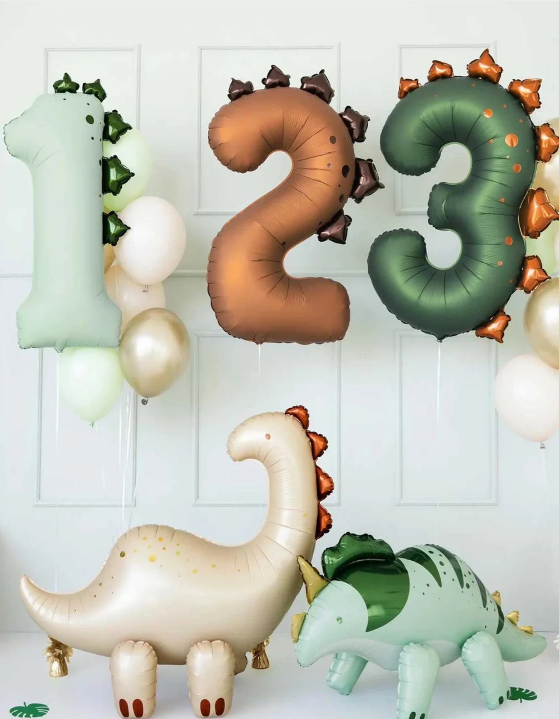 Dinosaur-themed birthday party decorations including large inflatable number balloons '1', '2', and '3' with dinosaur accents, plus two full dinosaur balloons, one cream/beige long-necked dinosaur and one mint green stegosaurus, set against a white paneled wall.