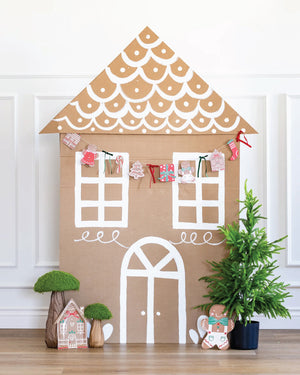 Decorative cardboard gingerbread house with a gingerbread Christmas garland with small plants and a teddy bear on a wooden floor.