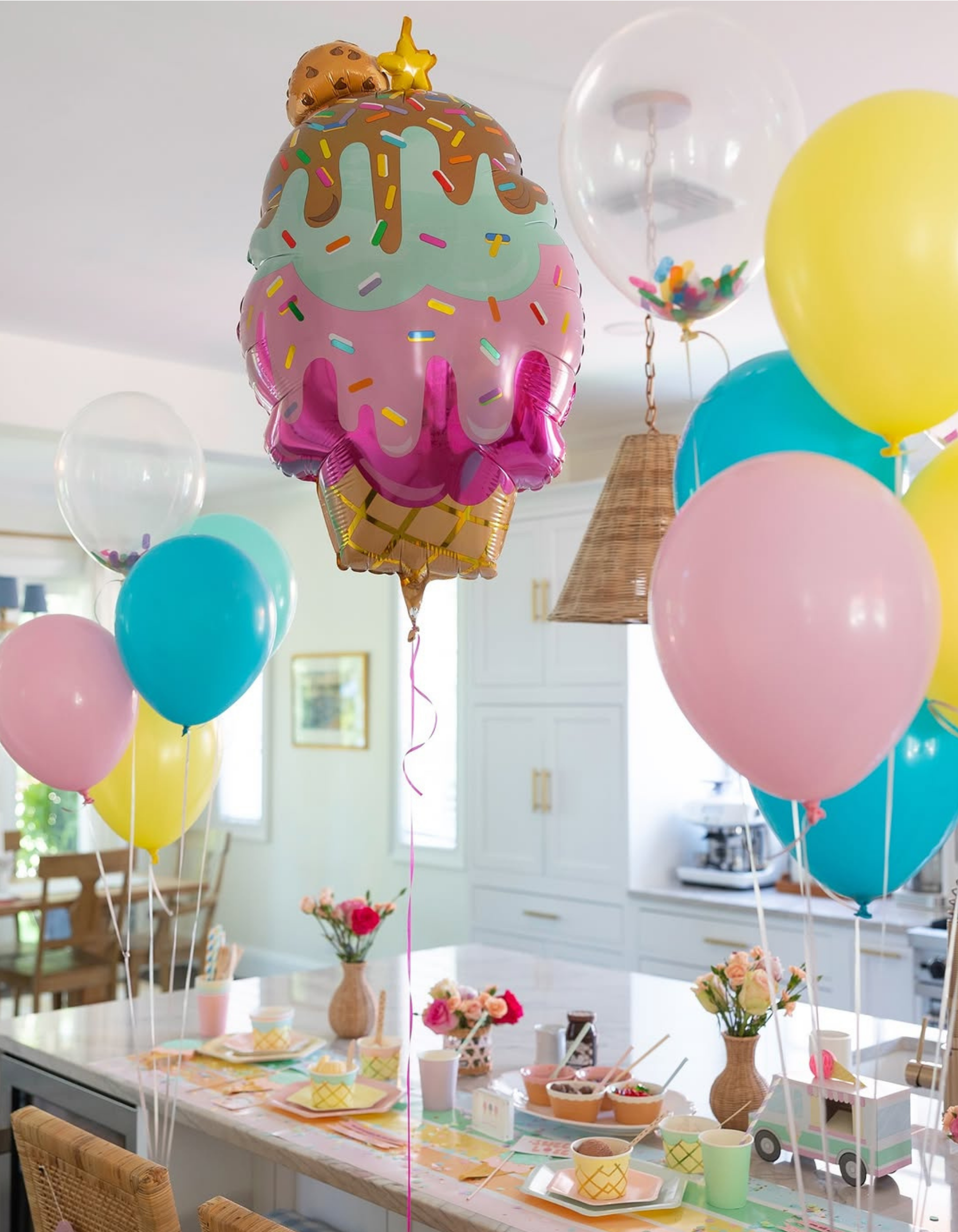 A party scene in a bright kitchen with a large balloon shaped like a frosted ice cream scoop floating above a decorated table. The balloon is pink and mint green with chocolate drizzle, sprinkles, and a small cookie on top.