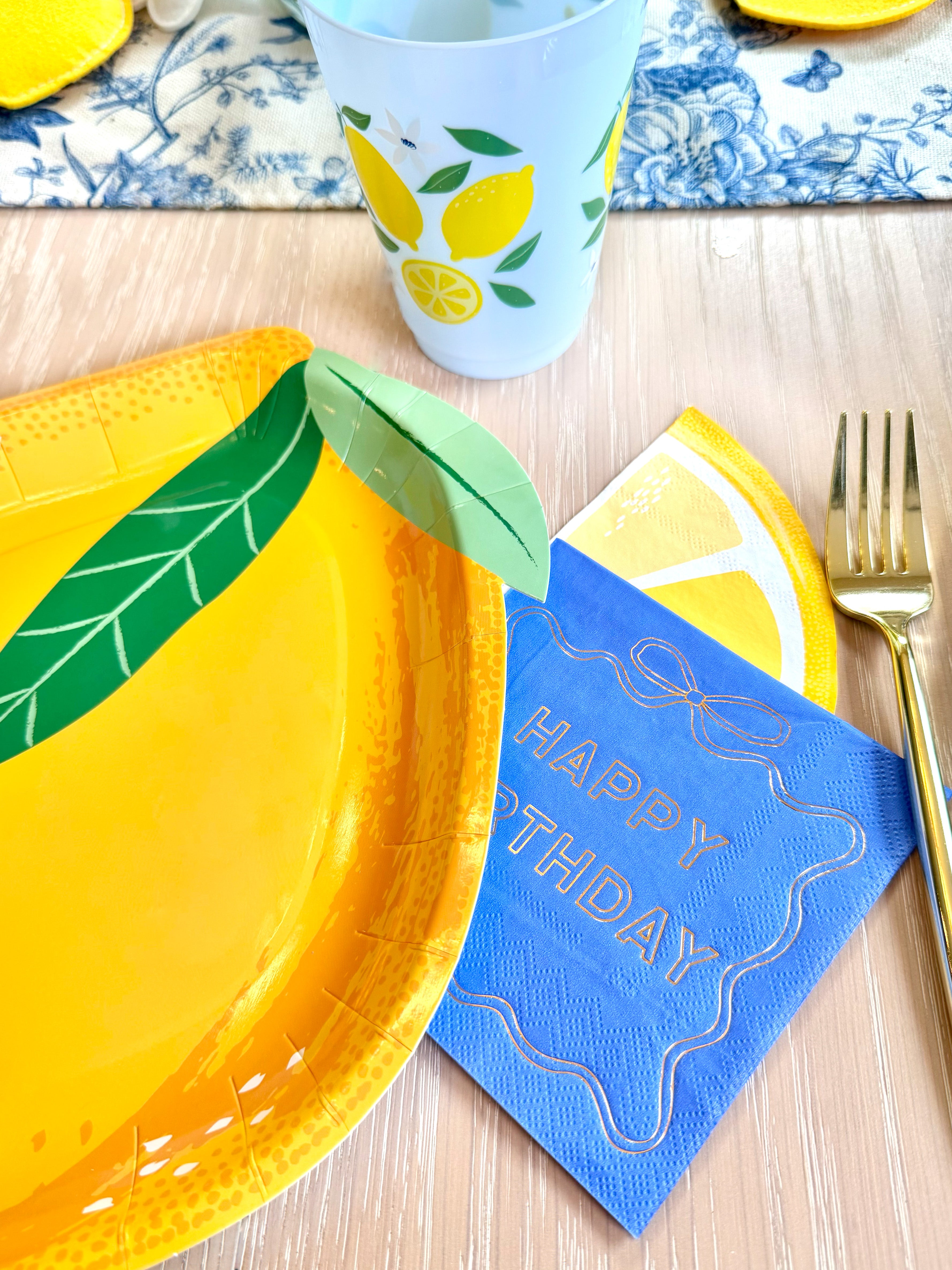 Lemon-shaped plate with blue “Happy Birthday” napkin, lemon-print cup, and gold fork on a light wood table.