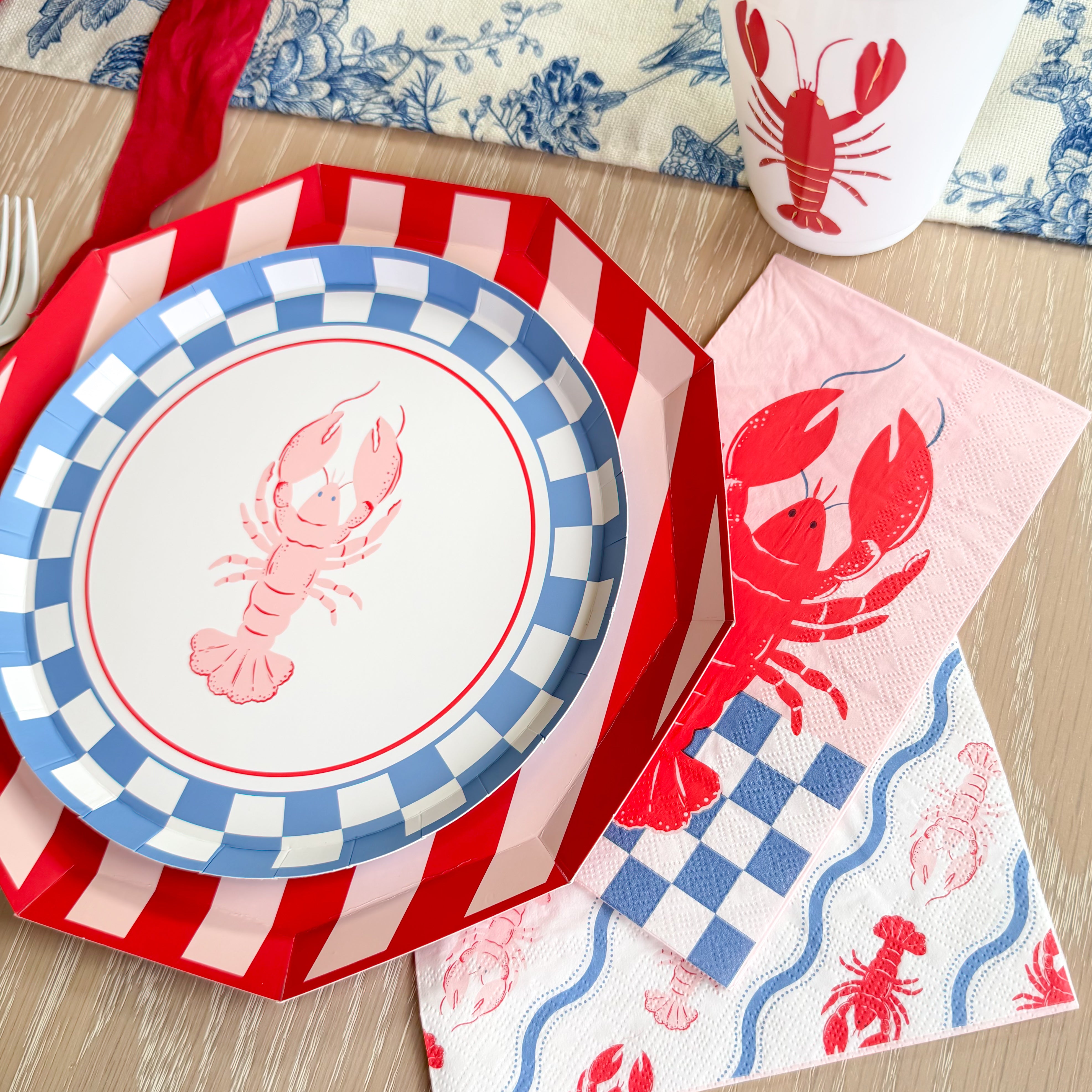 Lobster party table setting with floral centerpiece, striped plates, lobster cups, and blue toile runner.