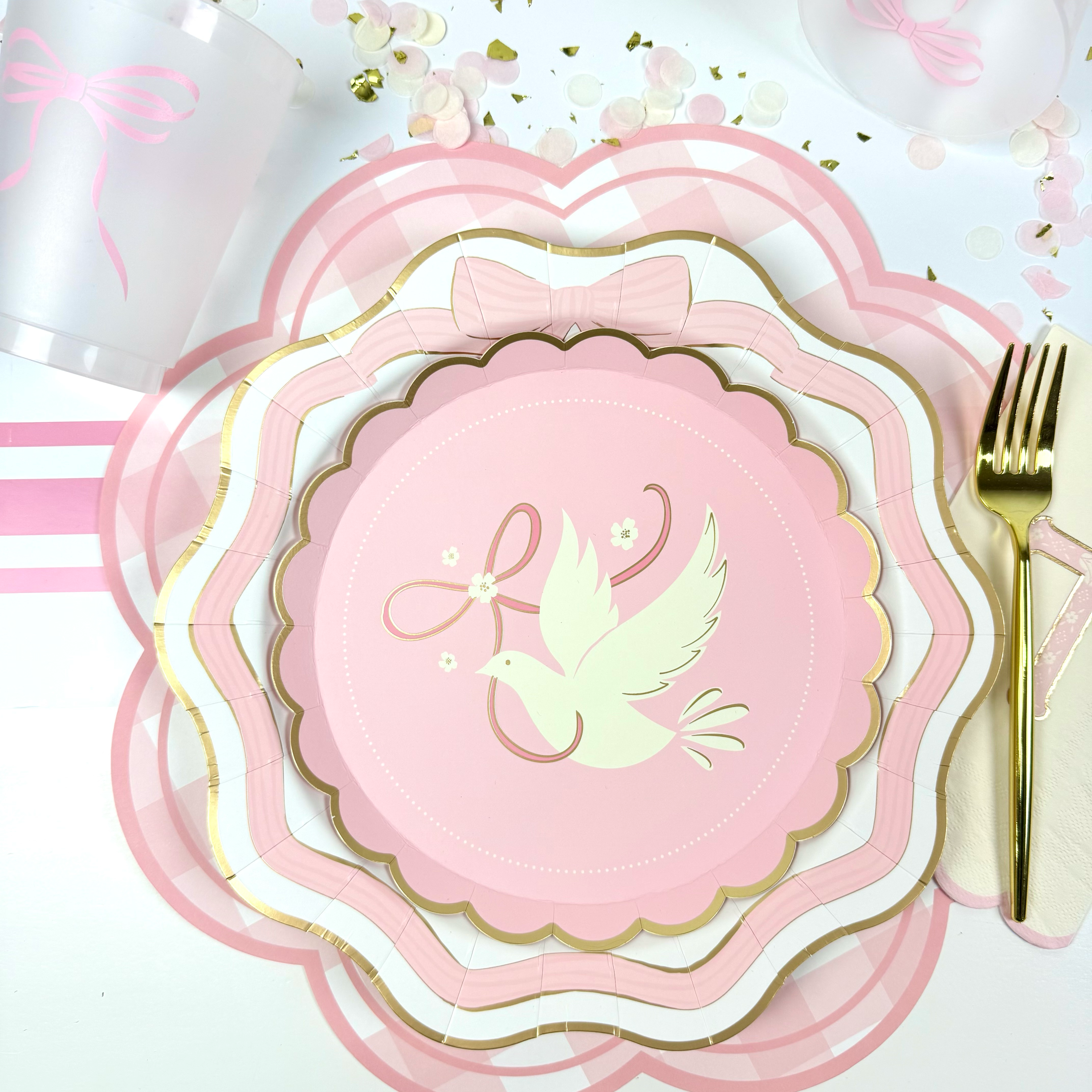 Pink dove-themed place setting with gold-trimmed scalloped plates, a gold fork, cross napkin, and scattered confetti.