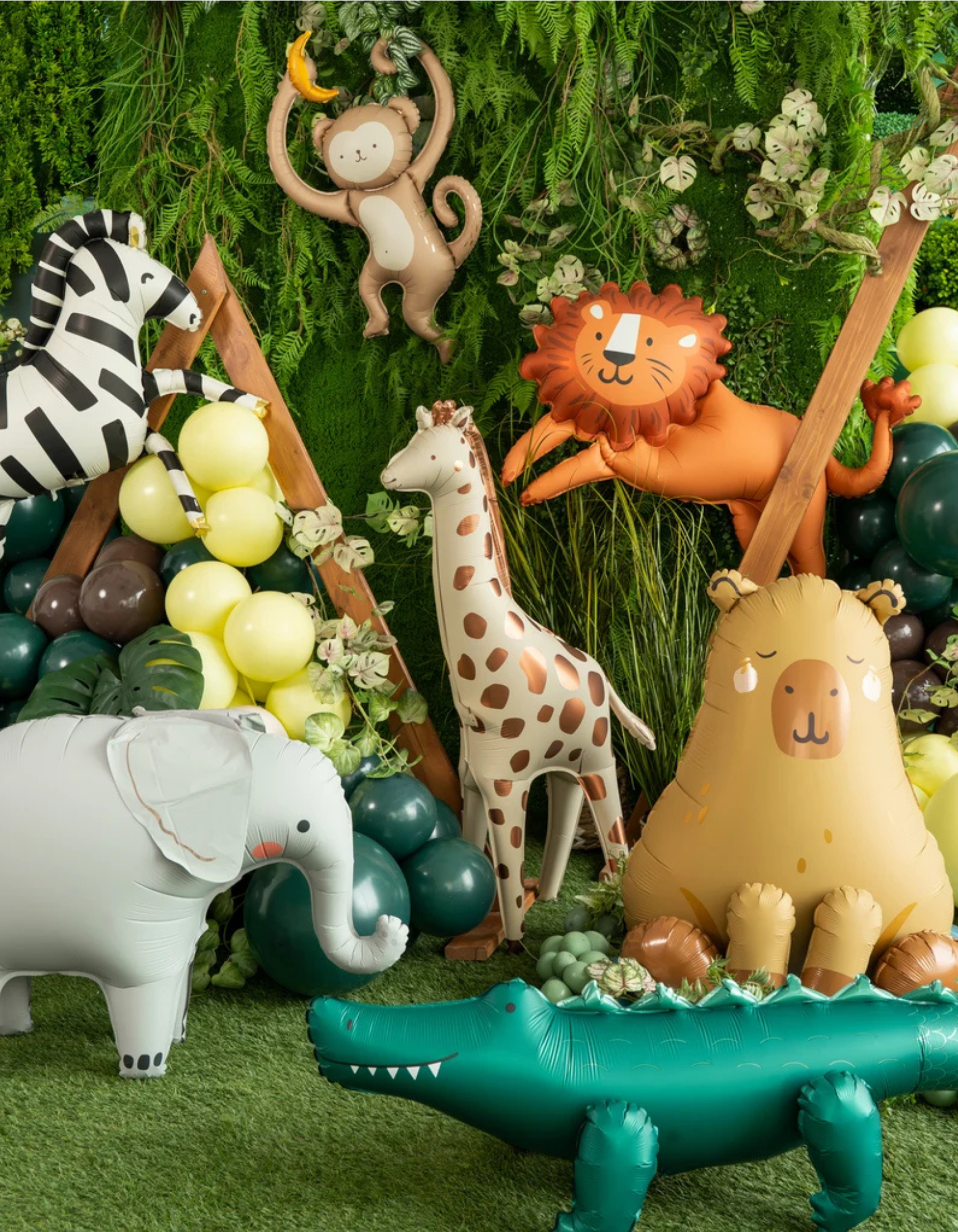 A styled jungle-themed party decoration display featuring multiple inflatable safari animal balloons including a zebra, giraffe, lion, elephant, capybara, and alligator, arranged against a green foliage backdrop with balloons and wooden props.