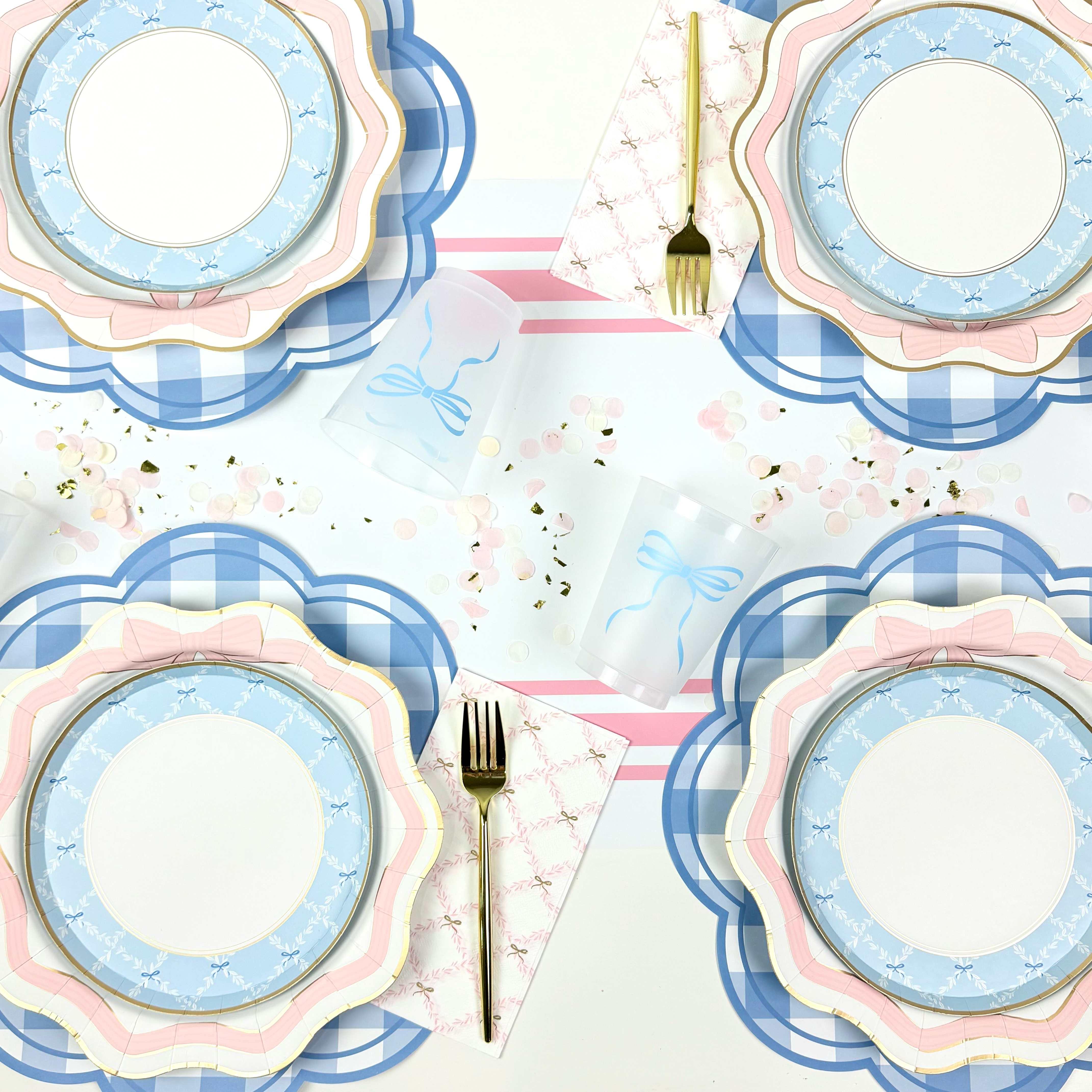 Overhead view of pink and blue bow-themed place settings with scalloped plates, gingham placemats, bow cups, and gold flatware.