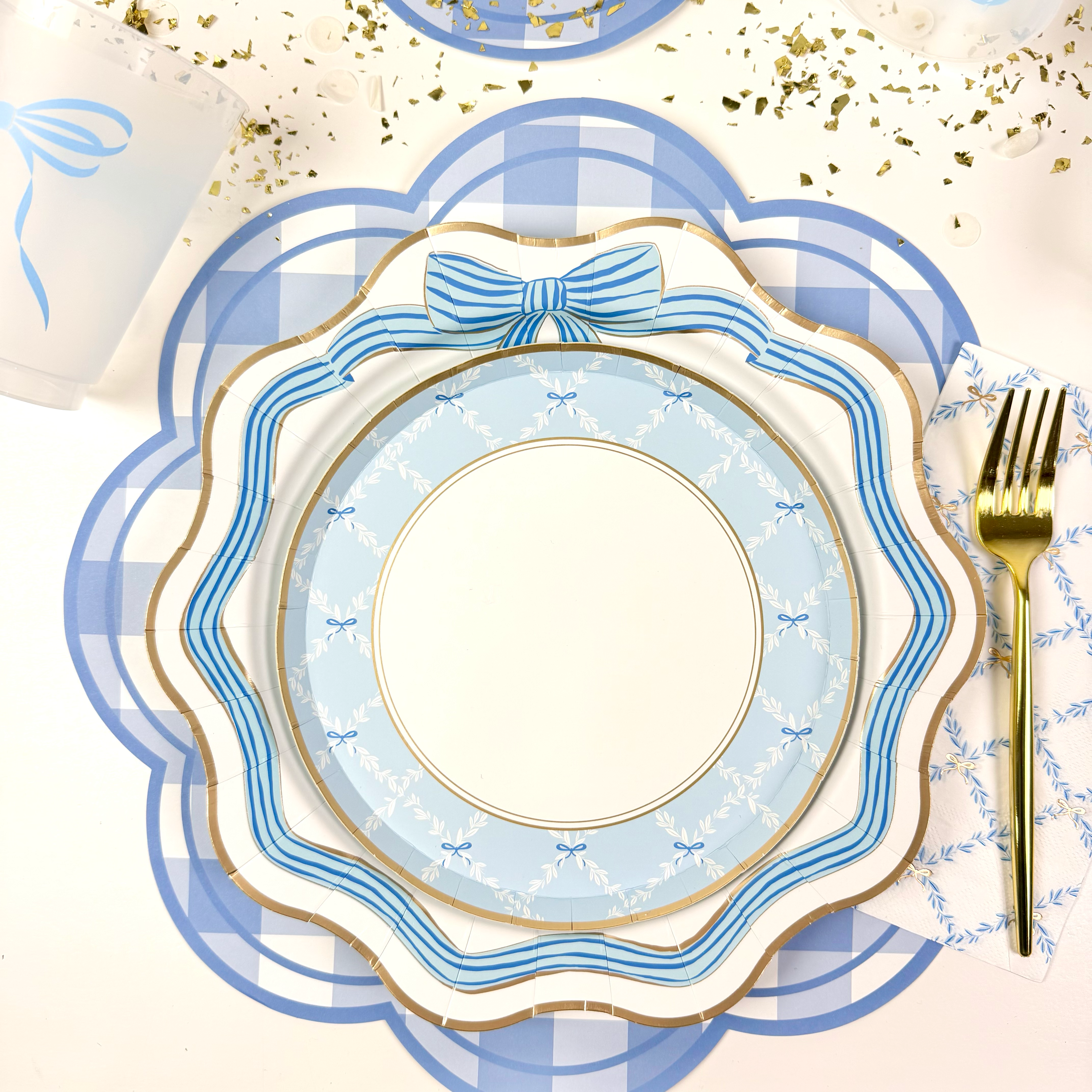 Close-up of blue and gold scalloped plates with bow accents layered on a blue gingham placemat.
