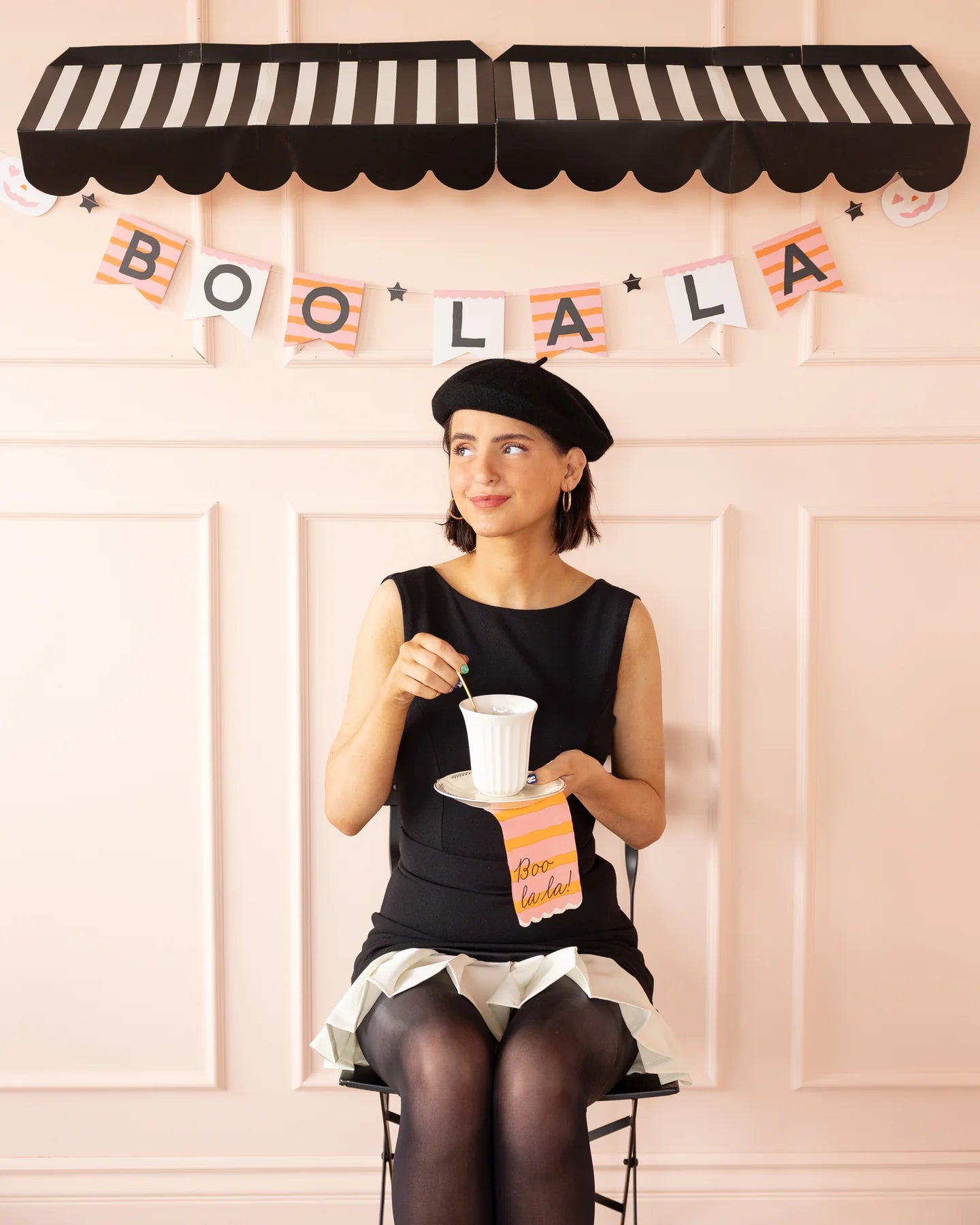 A woman sitting under a black and white striped awning with a Halloween 'BOO LA LA' banner for decorations.