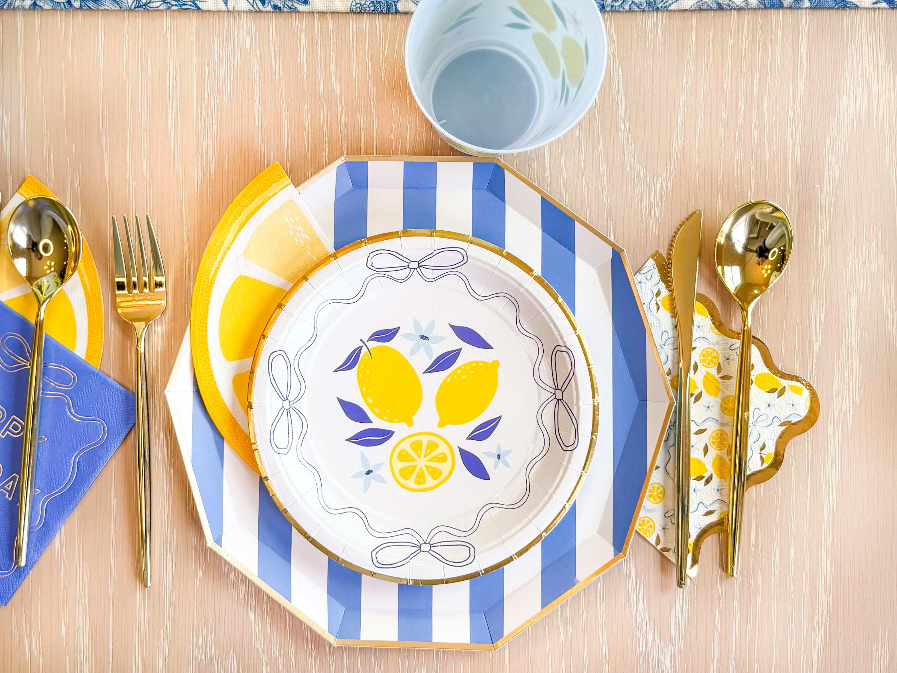 Coastal lemon place setting with striped charger, lemon plate, lemon slice accent, and gold flatware.