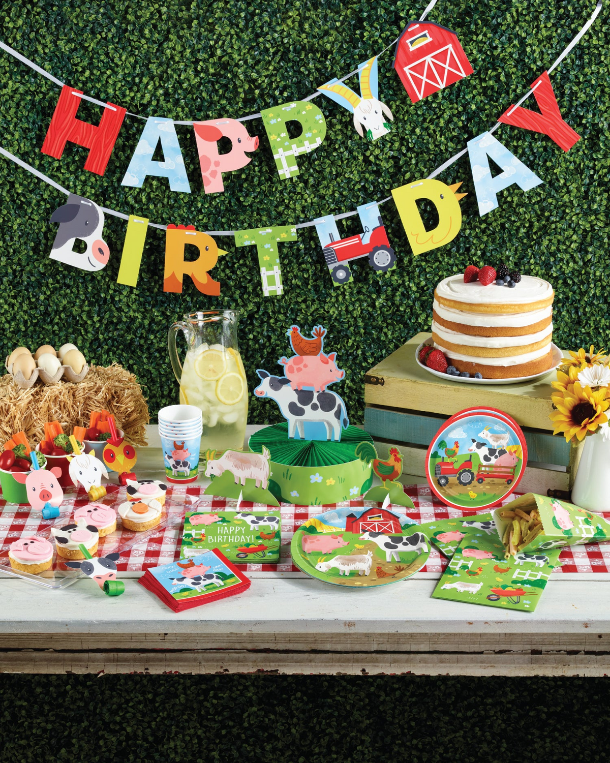 Farm-themed birthday party setup with cake, cupcakes, and decorations against a green hedge background.