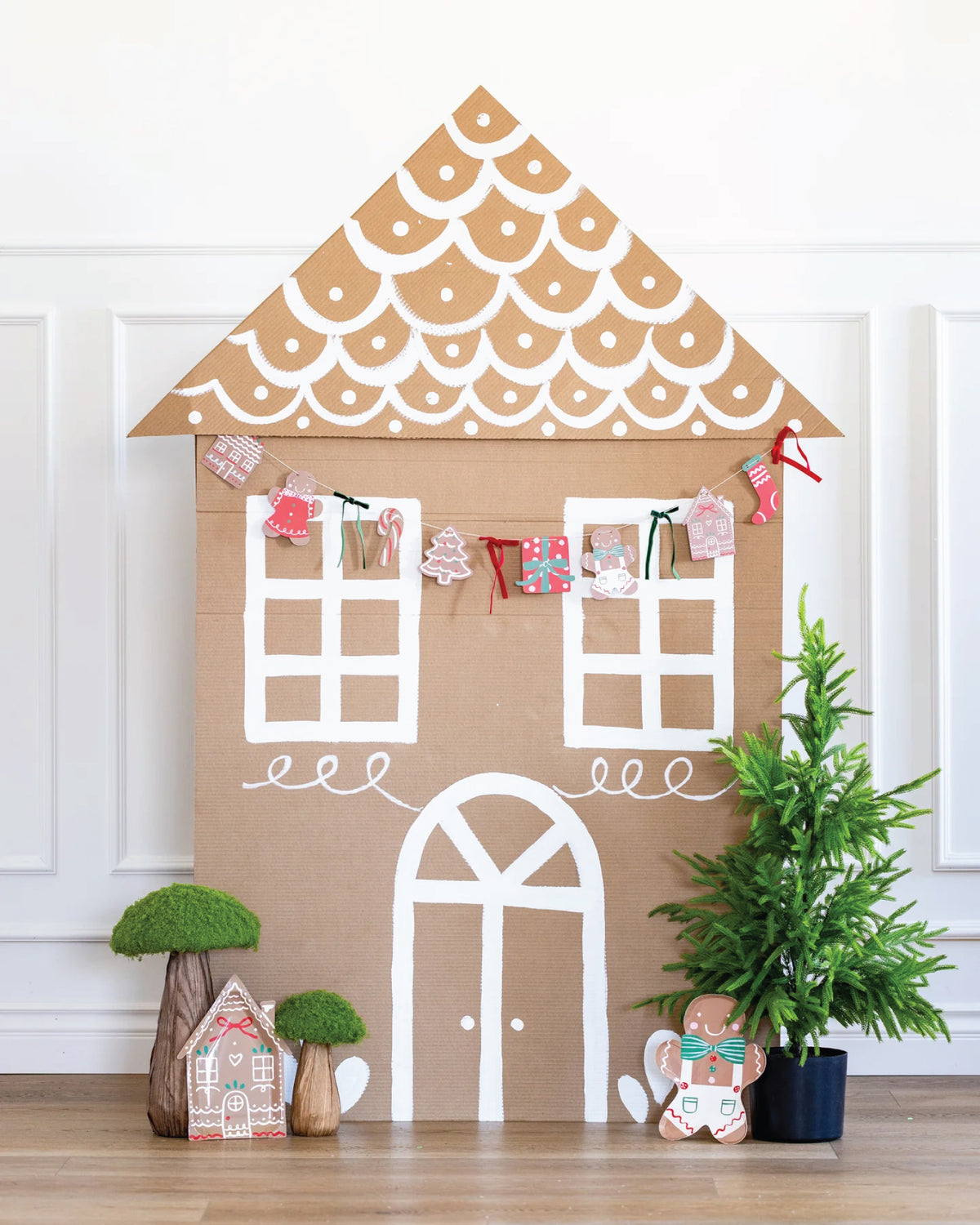Decorative cardboard gingerbread house with a gingerbread Christmas garland with small plants and a teddy bear on a wooden floor.