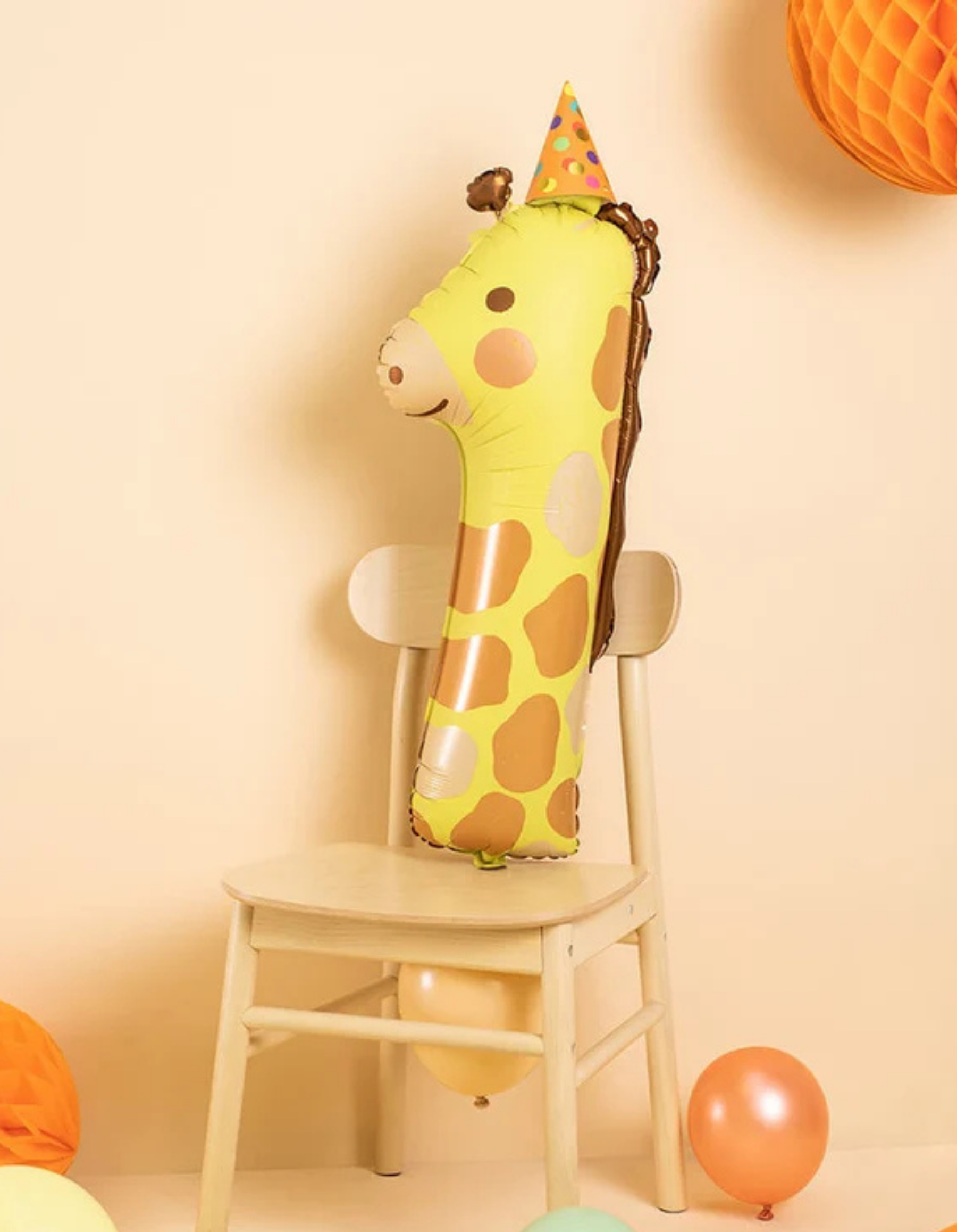 A yellow and brown giraffe-shaped foil number one balloon wearing a party hat, displayed on a light wood chair against a warm beige wall