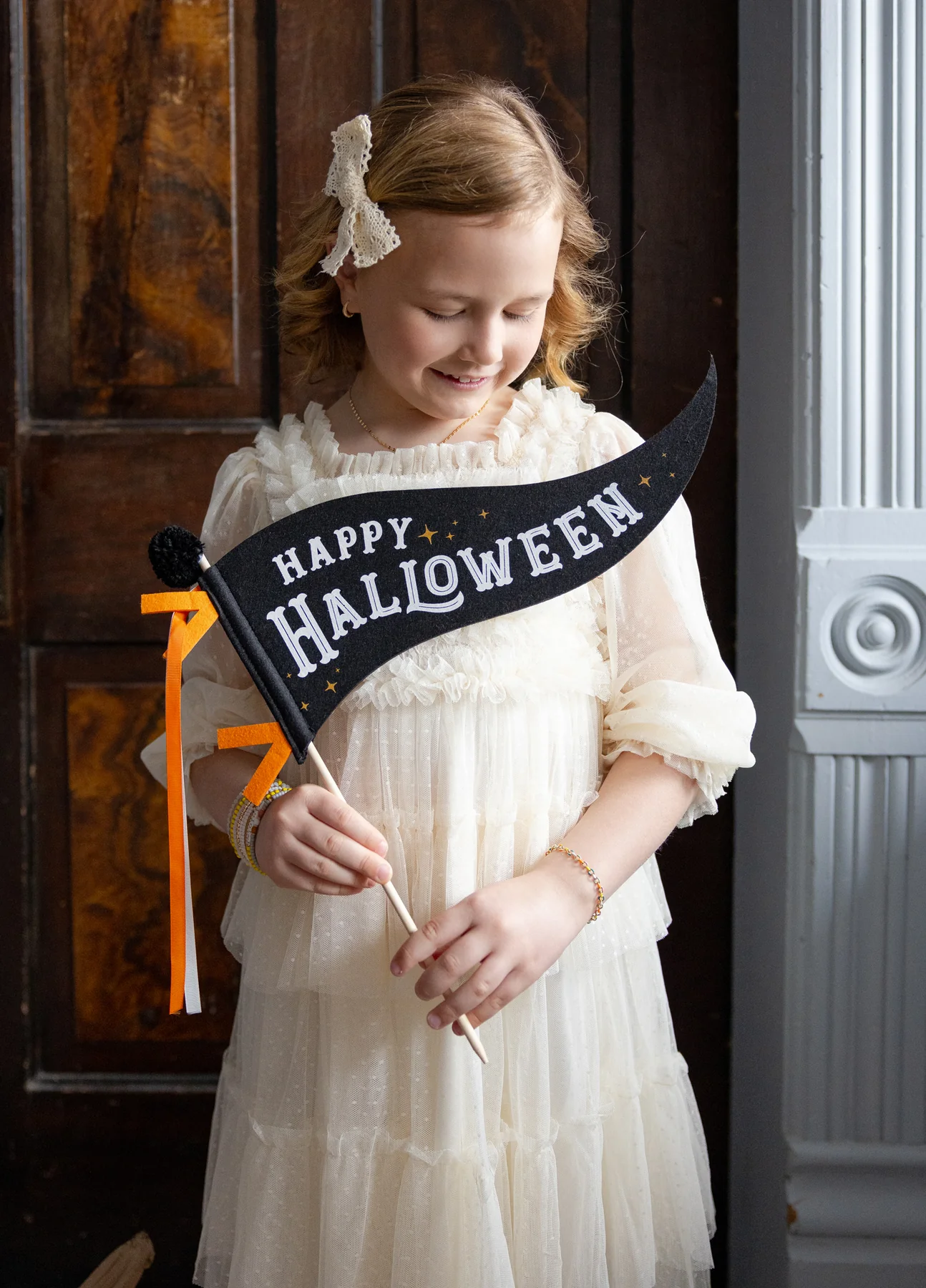 Young girl in a white dress holding a 'Happy Halloween' felt pennant.