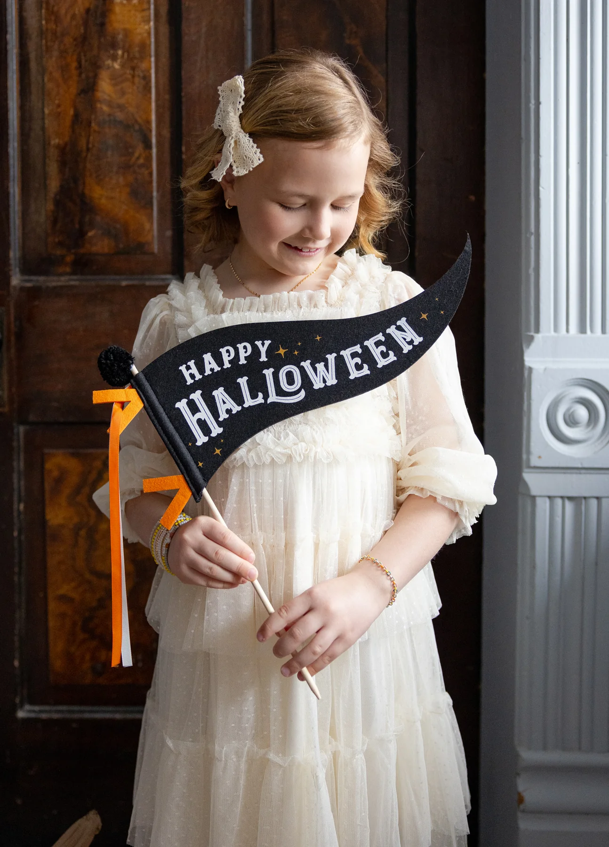 Young girl in a white dress holding a &#39;Happy Halloween&#39; felt pennant.