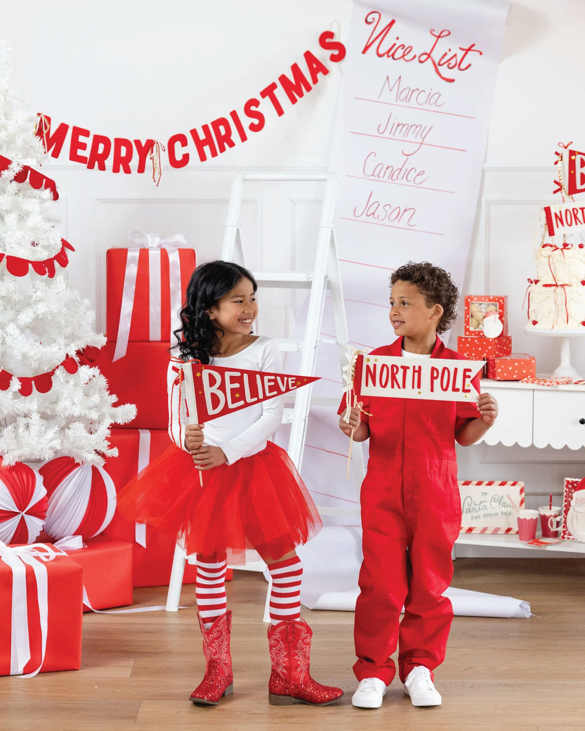 Two children in Christmas-themed outfits holding Believe & North Pole Felt Pennant Flags in a decorated room with a Christmas tree and presents.