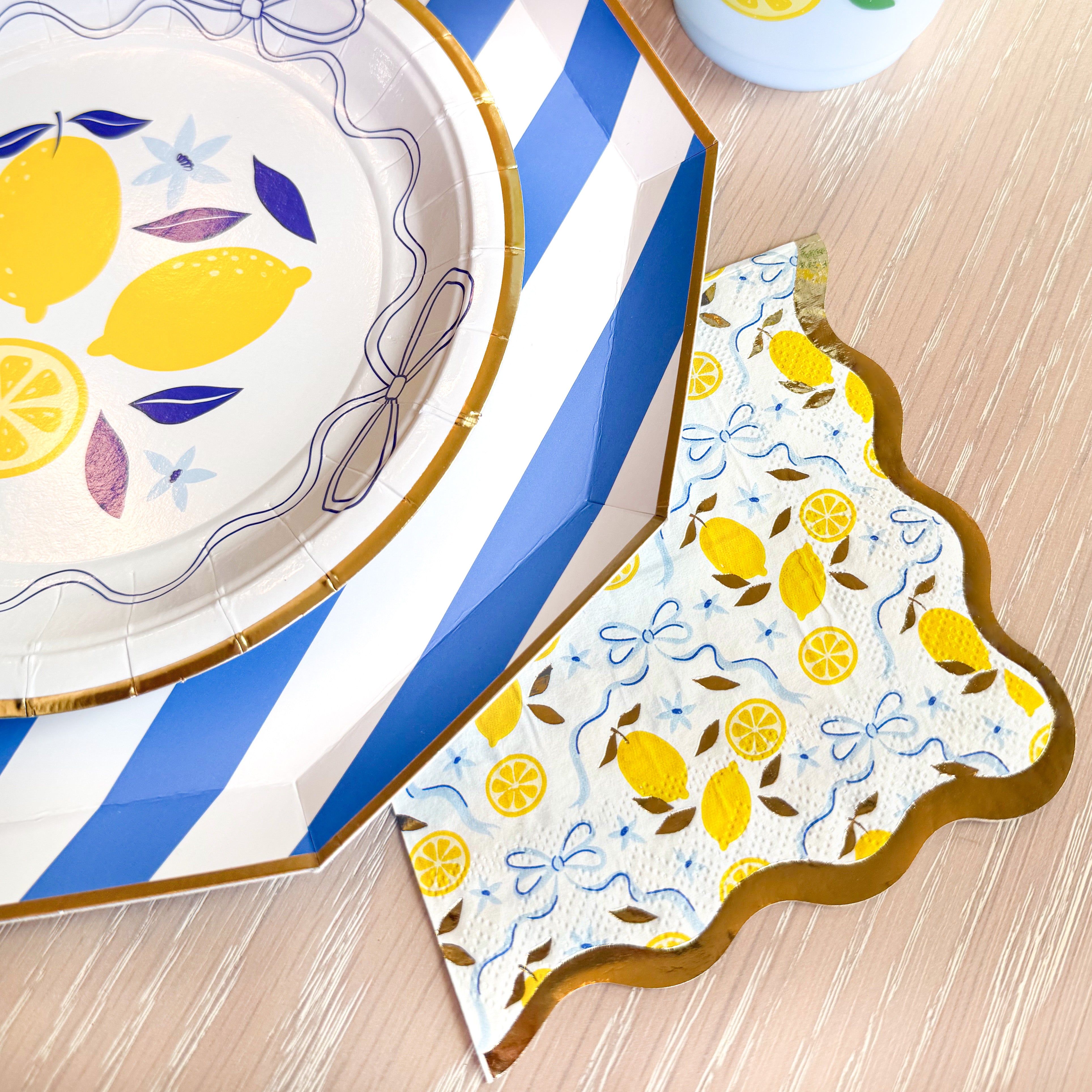Lemon-print cocktail napkin with gold scalloped edge beside a blue striped charger and lemon-themed plate.