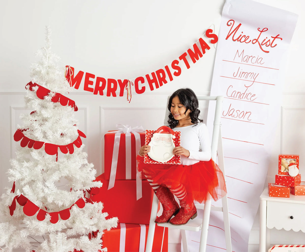 Child in festive Christmas setting holding a Santa Claus Paper Lunch Plates surrounded by a white tree, presents, and a 'Merry Christmas' banner.