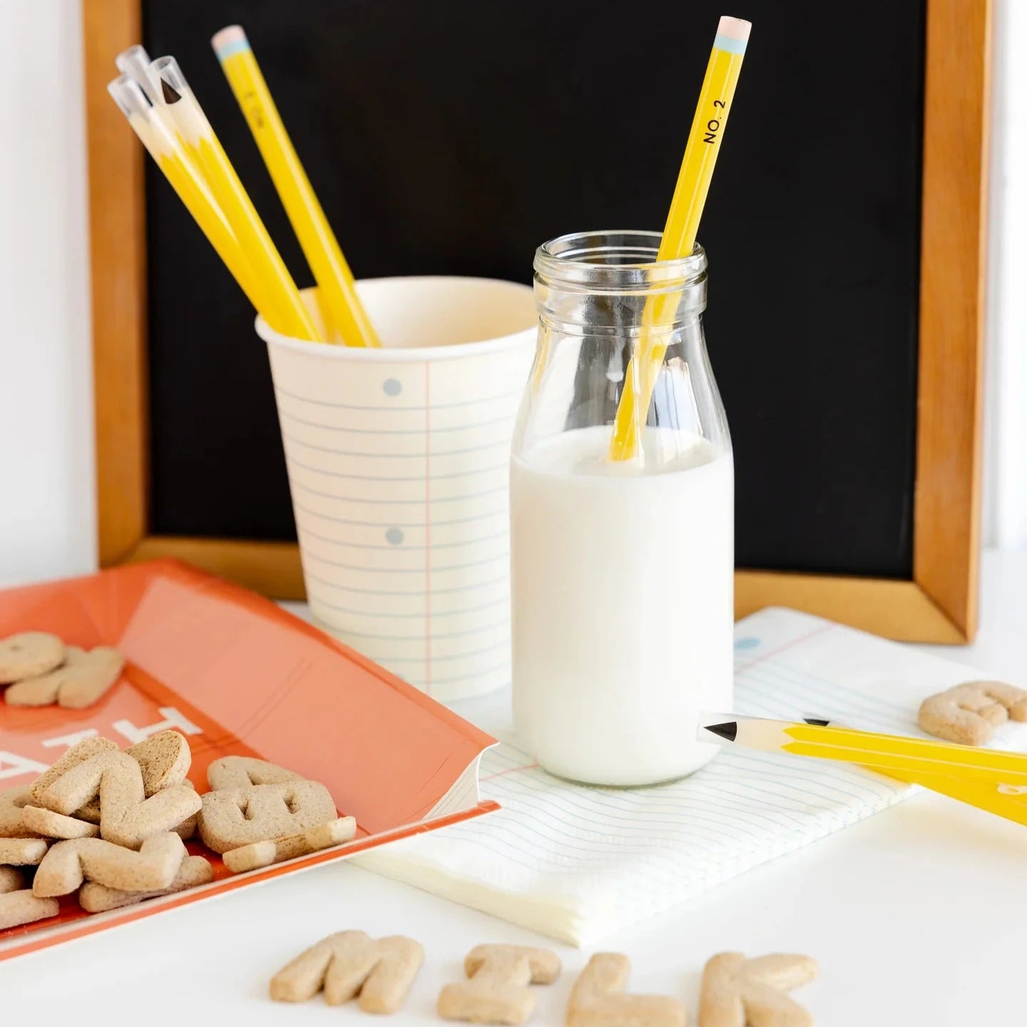 Milk bottle with a straw, pencils, and alphabet cookies on a table.