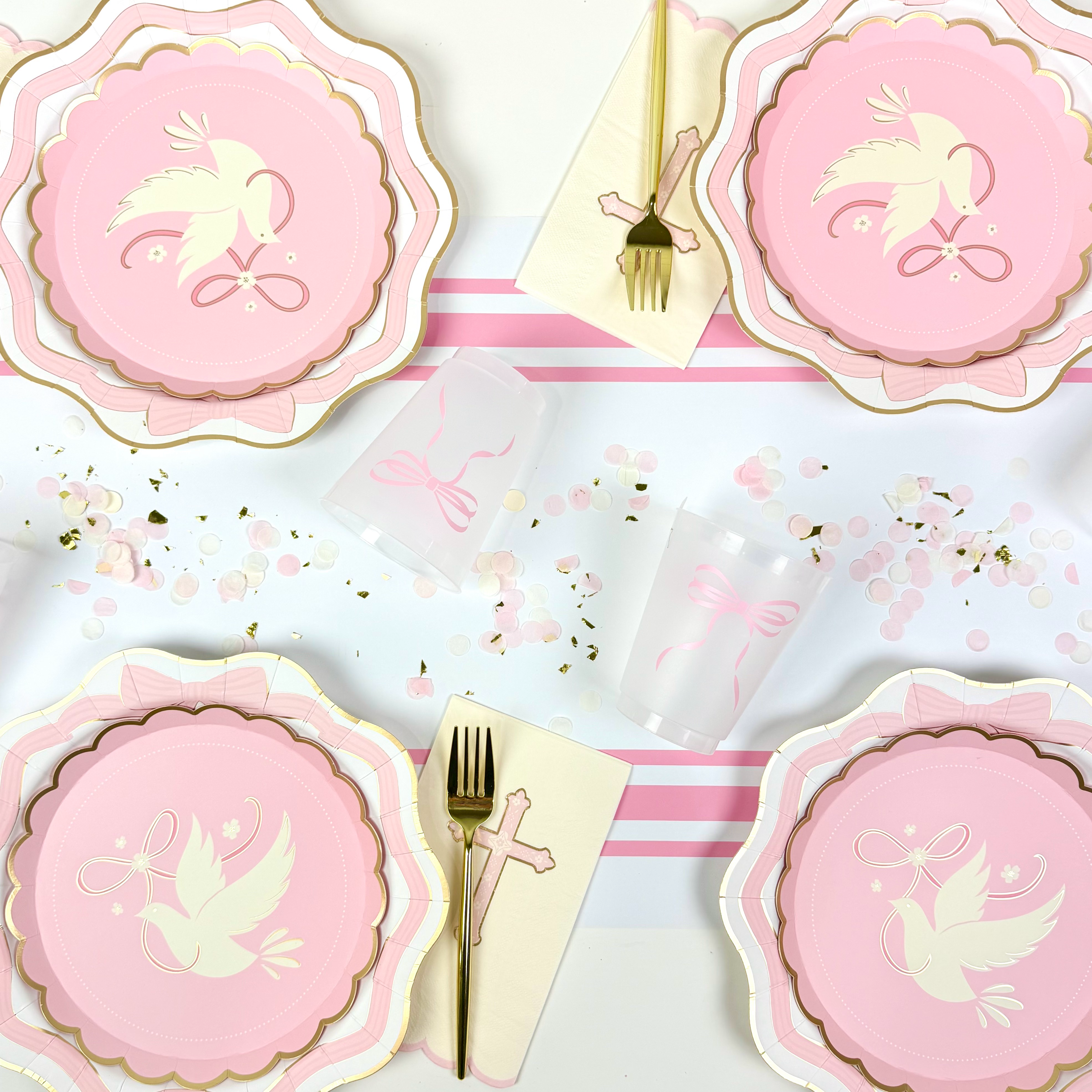 Overhead view of a pink baptism table setting with dove plates, bow cups, gold forks, cross napkins, and pastel confetti.
