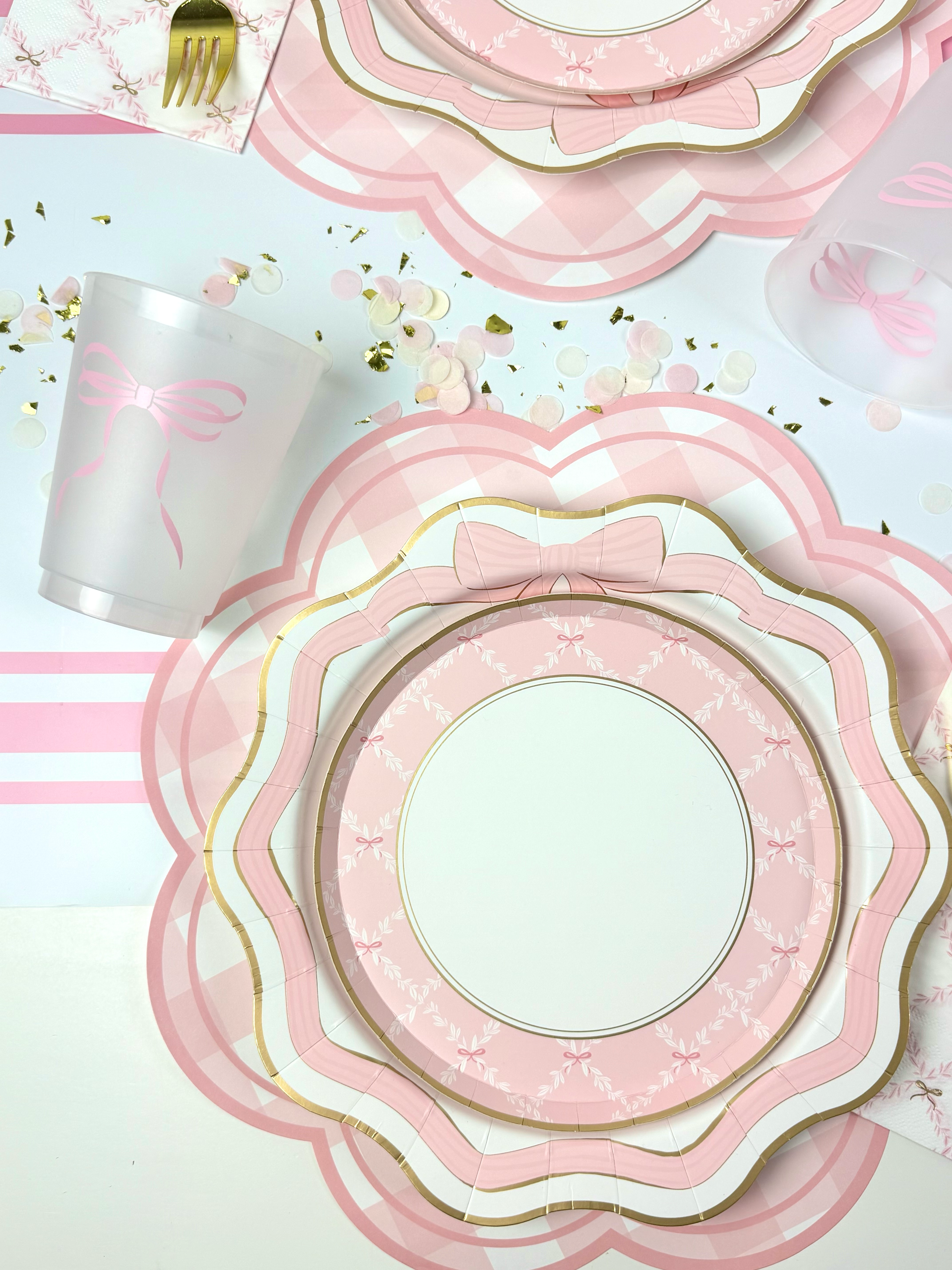 Pink bow-themed place setting with scalloped gold-trimmed plates, gingham placemat, bow cup, and scattered confetti.
